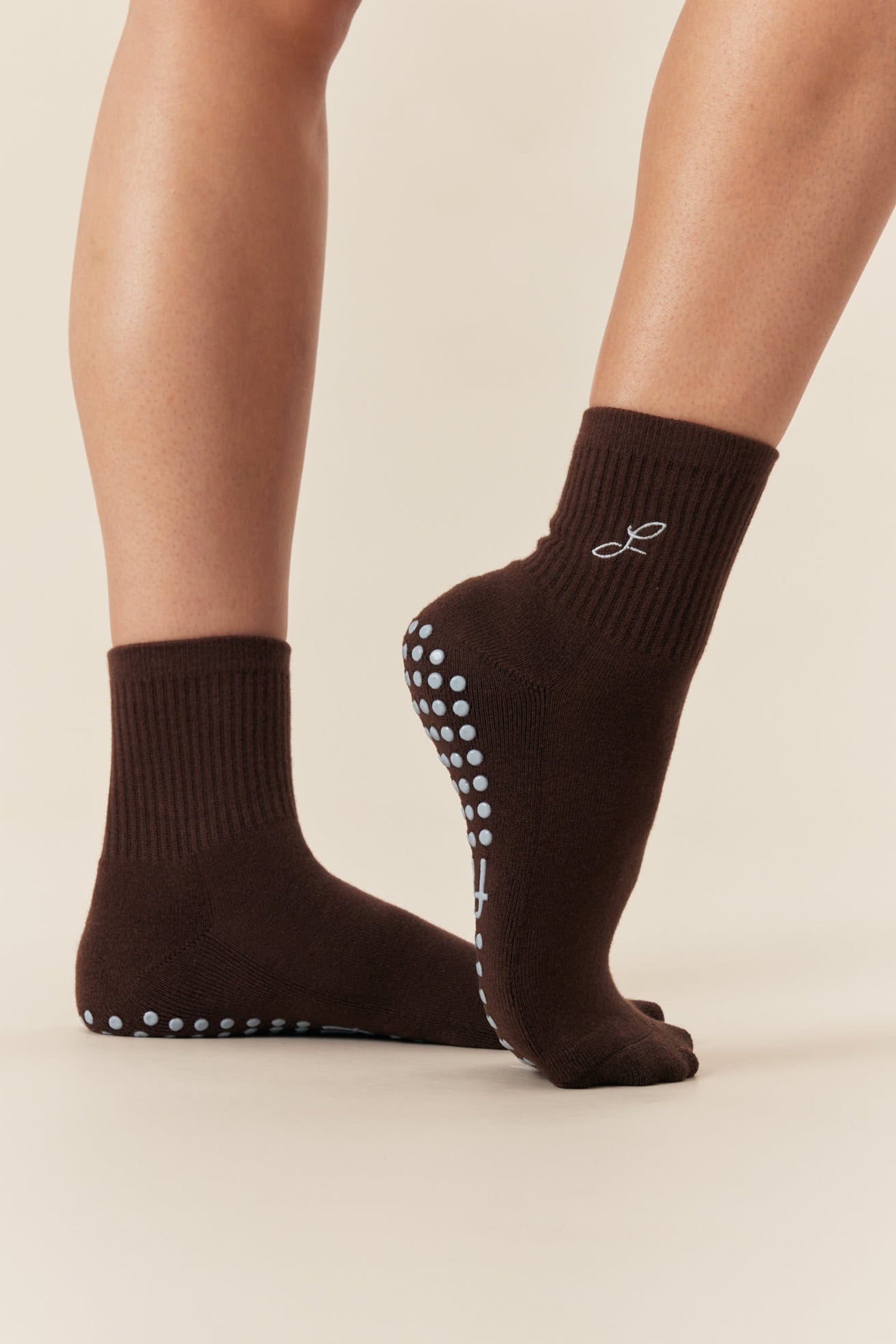 A pair of dark brown ankle socks worn on a model's feet, featuring BLUE grip dots on the sole and heel and a small FIRMA symbol on the side.