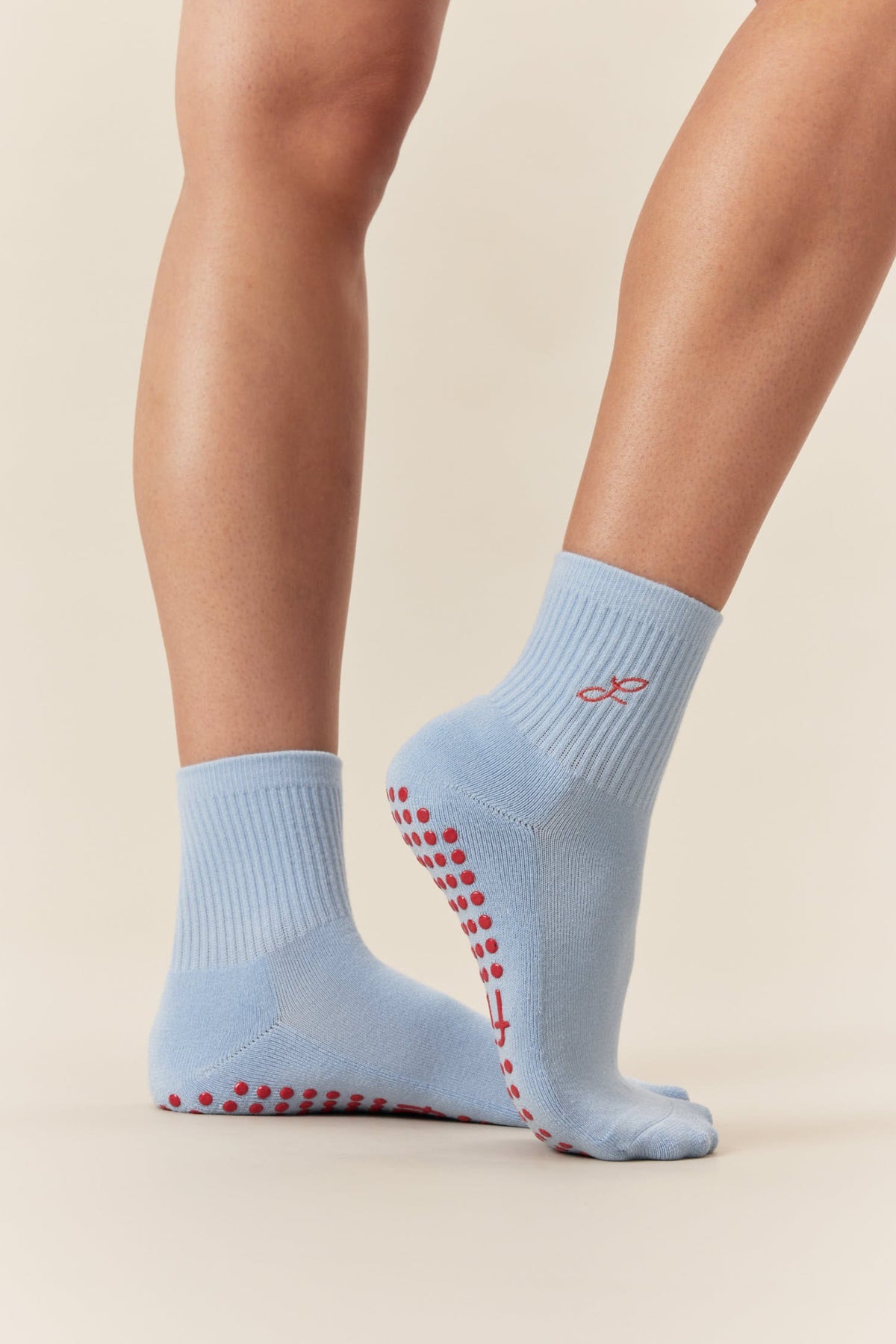 A pair of light blue ankle socks with red grip dots on the sole and a small red logo on the side, shown on a model's feet against a warm beige background.